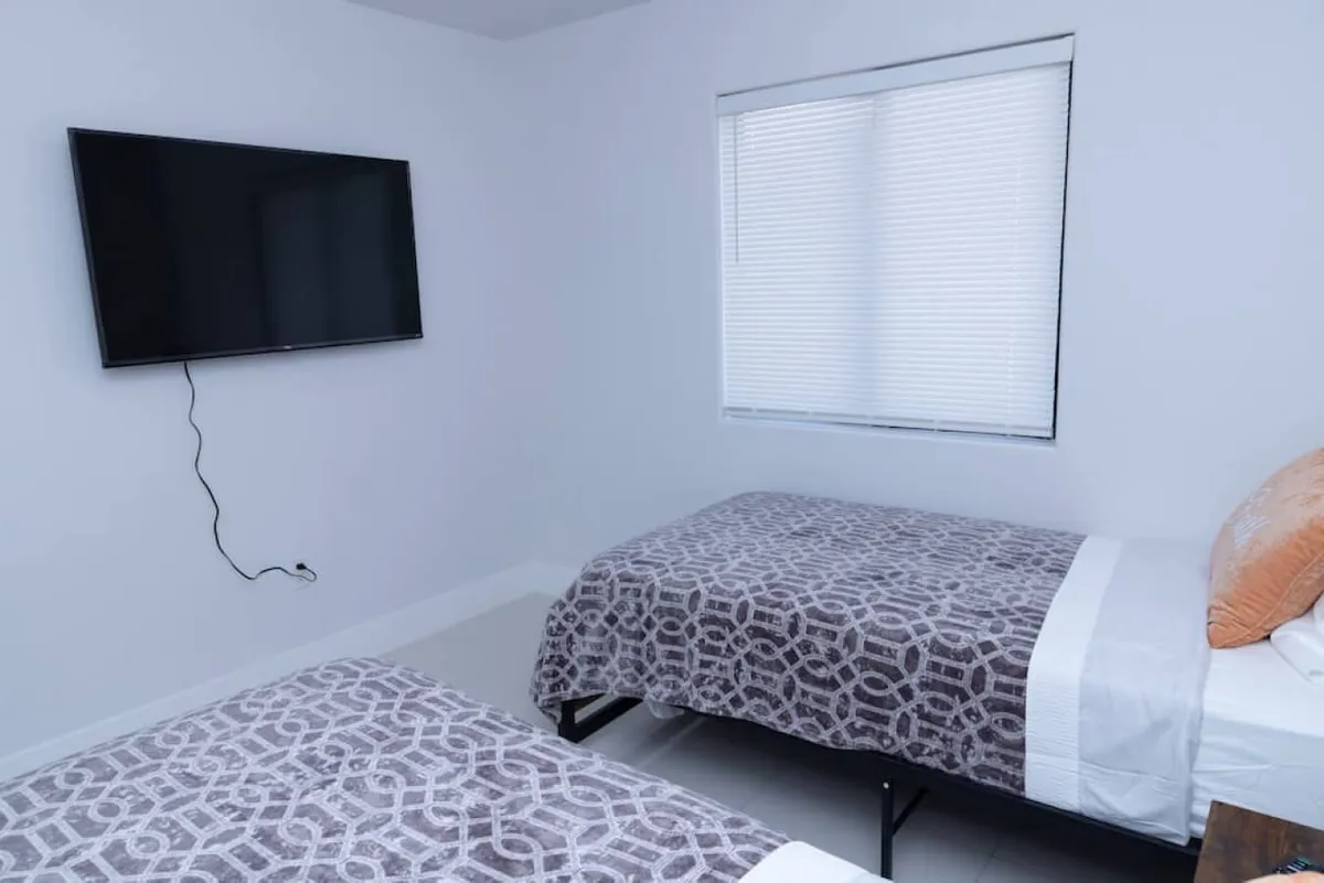 A bright, inviting bedroom featuring two beds, a flat-screen TV, and ample natural light streaming through the window. The image conveys comfort and modern amenities, with space that feels clean and ready for fresh starts after using the laundry facilities.