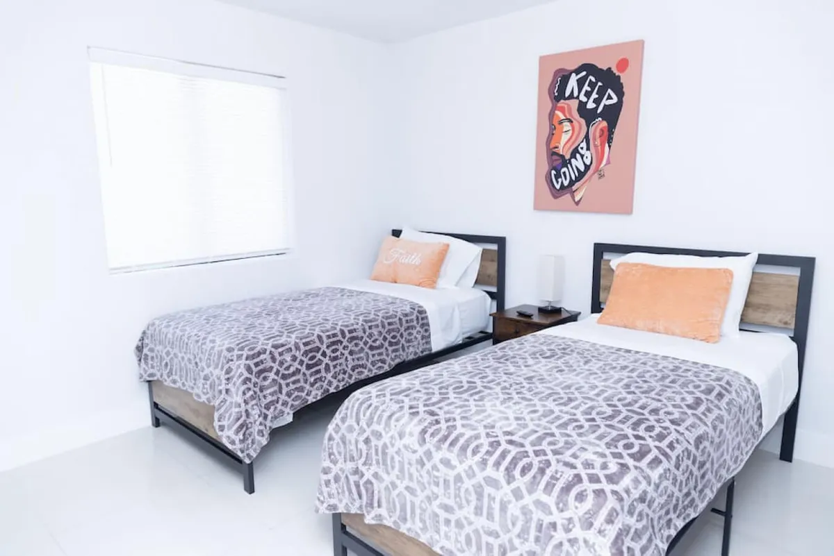 A bright, uplifting bedroom with two beds dressed in patterned linens, motivational wall art above the headboards, and a nightstand with a lamp. The ambiance is both serene and inviting, suitable for families or groups.