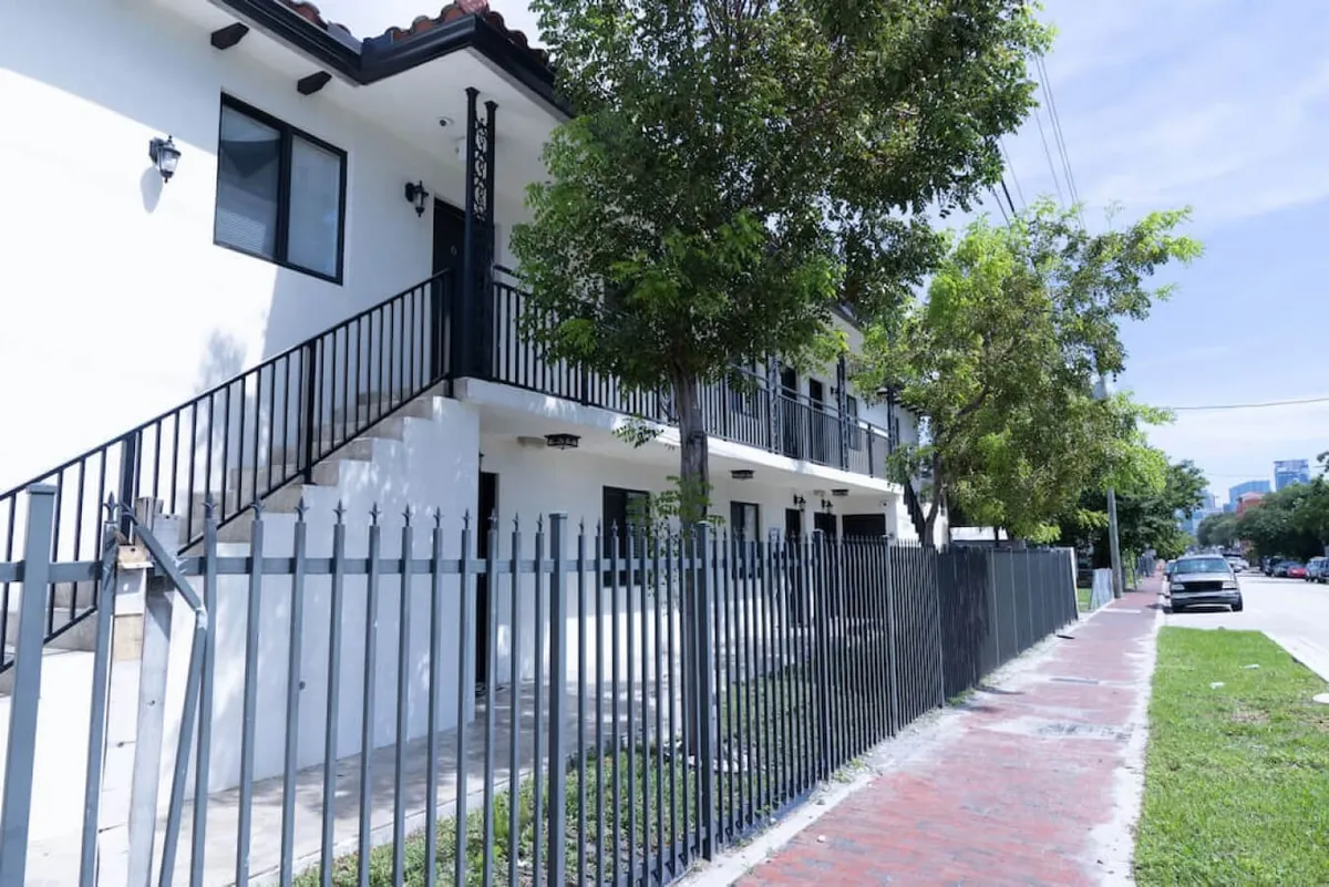 An exterior view showcasing the staircase leading up to the apartment, surrounded by lush green trees and a secure fence. The parking area is visible nearby, hinting at the ease of access from car to door. The space feels welcoming, safe, and convenient for arrivals.