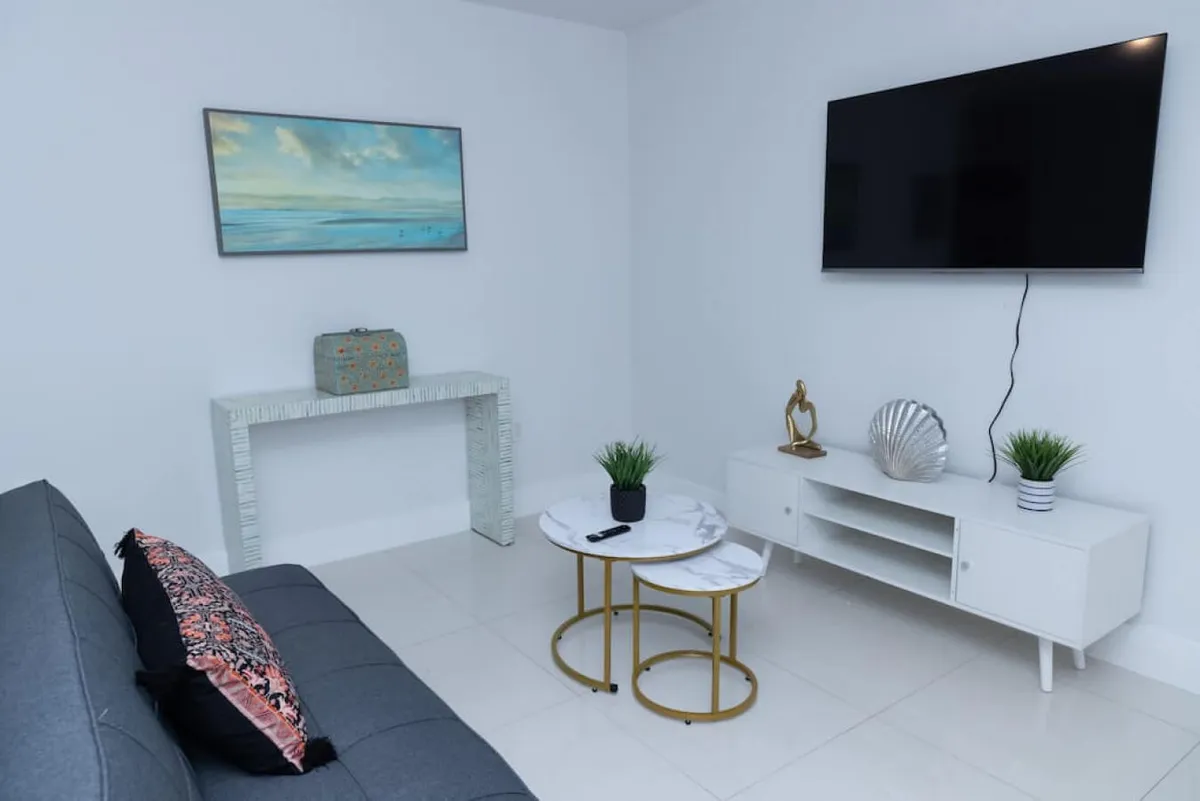 The image showcases a sleek, modern living area with a plush black sofa accented by decorative pillows, stylish nesting coffee tables with marble tops and gold frames, a wall-mounted flat screen TV, and a white TV stand adorned with tasteful decor. The space exudes comfort and contemporary design, creating an inviting atmosphere for relaxation or socializing.