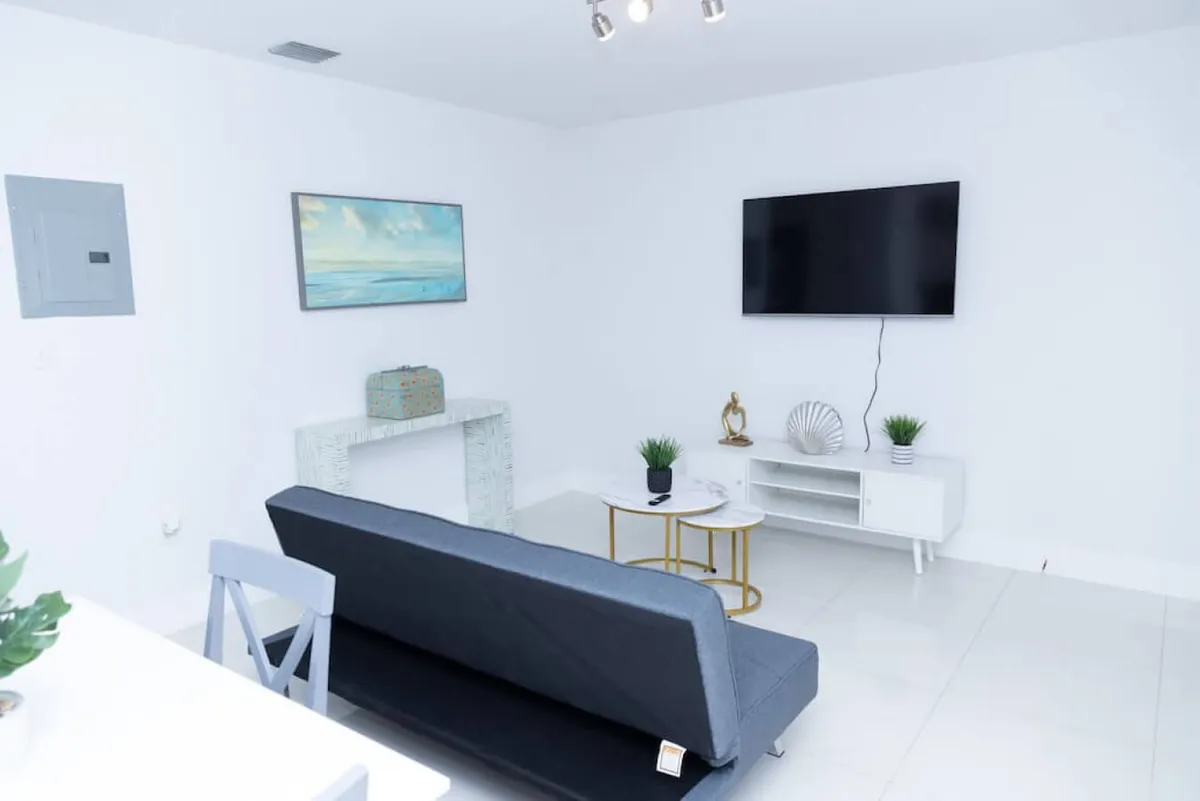 This image features a calm and inviting living room area, showcasing a wall-mounted flat screen TV, a dark grey sofa bed, and a white modern TV console topped with decorative items. An abstract ocean-themed painting adds a touch of local flavor and color to the space. The layout is open and comfortable, creating a welcoming first impression for guests entering the apartment.