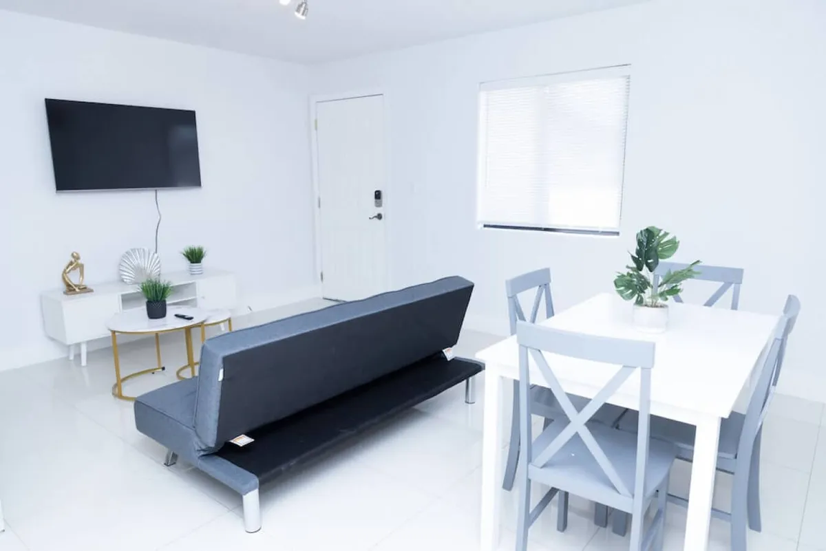 This image features a fold-out sofa bed in the living area, a wall-mounted TV, and a white dining table with four chairs. Modern minimalist decor accents the space, and the setup is ideal for both relaxation and hosting guests.