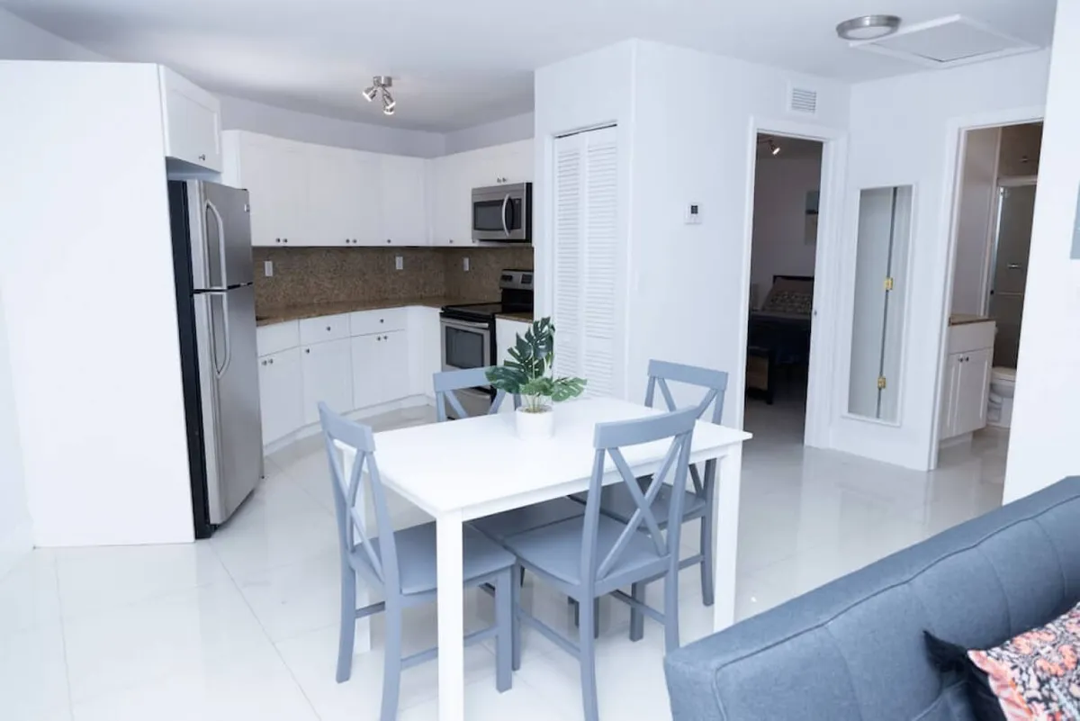 This image presents a bright, welcoming open-plan living space that seamlessly connects the living room, dining area, and modern kitchen. The white dining table with blue chairs sits adjacent to a gray sofa, while the modern kitchen with stainless steel appliances is visible in the background. Natural light pours in, highlighting the openness and functionality of the space.