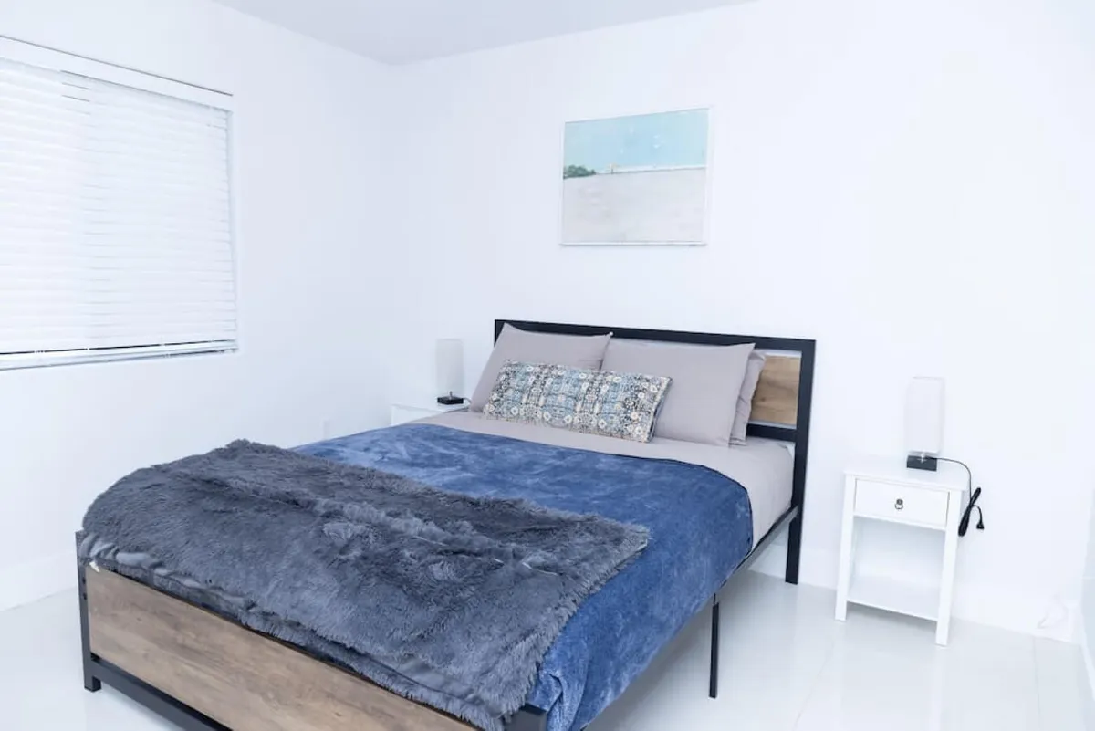 A serene bedroom featuring a queen-sized bed dressed in soft, layered linens. Two minimalist bedside tables with lamps flank the bed, creating a cozy atmosphere. A window with blinds allows for natural light, while abstract wall art above the bed adds a touch of personality.