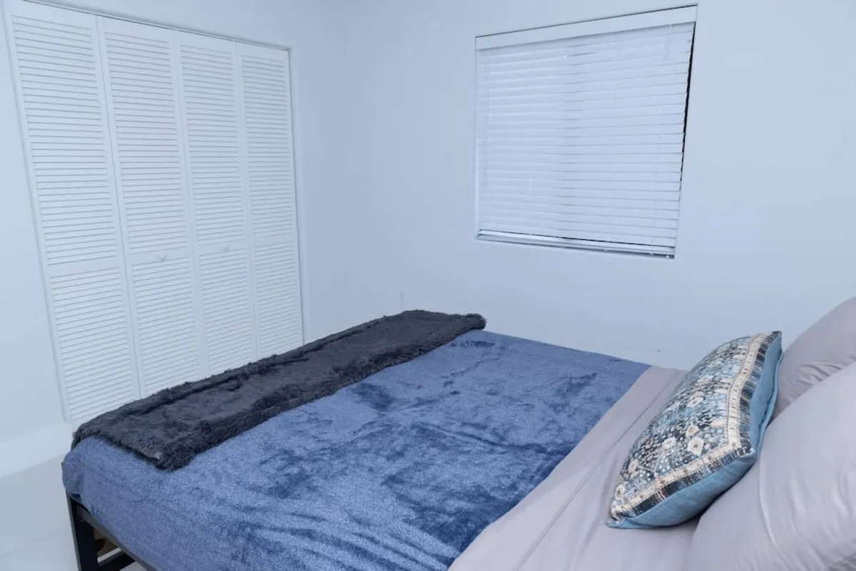 A quiet, inviting bedroom with a bed dressed in blue and gray bedding and a decorative pillow. White closet doors provide convenient storage, and a window with closed blinds ensures privacy and calm. The room exudes restful simplicity, perfect for recharging.