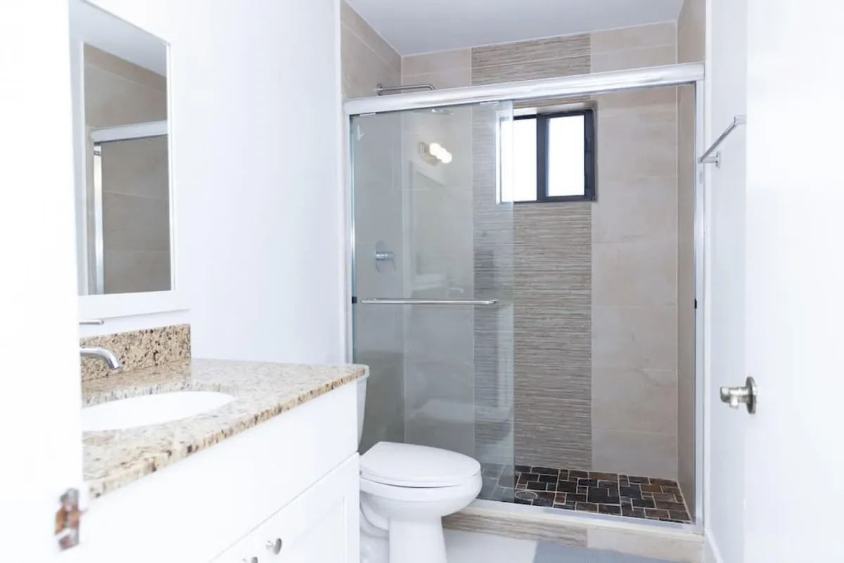 A modern bathroom featuring a glass sliding shower door, granite countertop with sink, toilet, and a small window above the shower. The space is bright and functional, ideal for morning routines or refreshing after a day out.